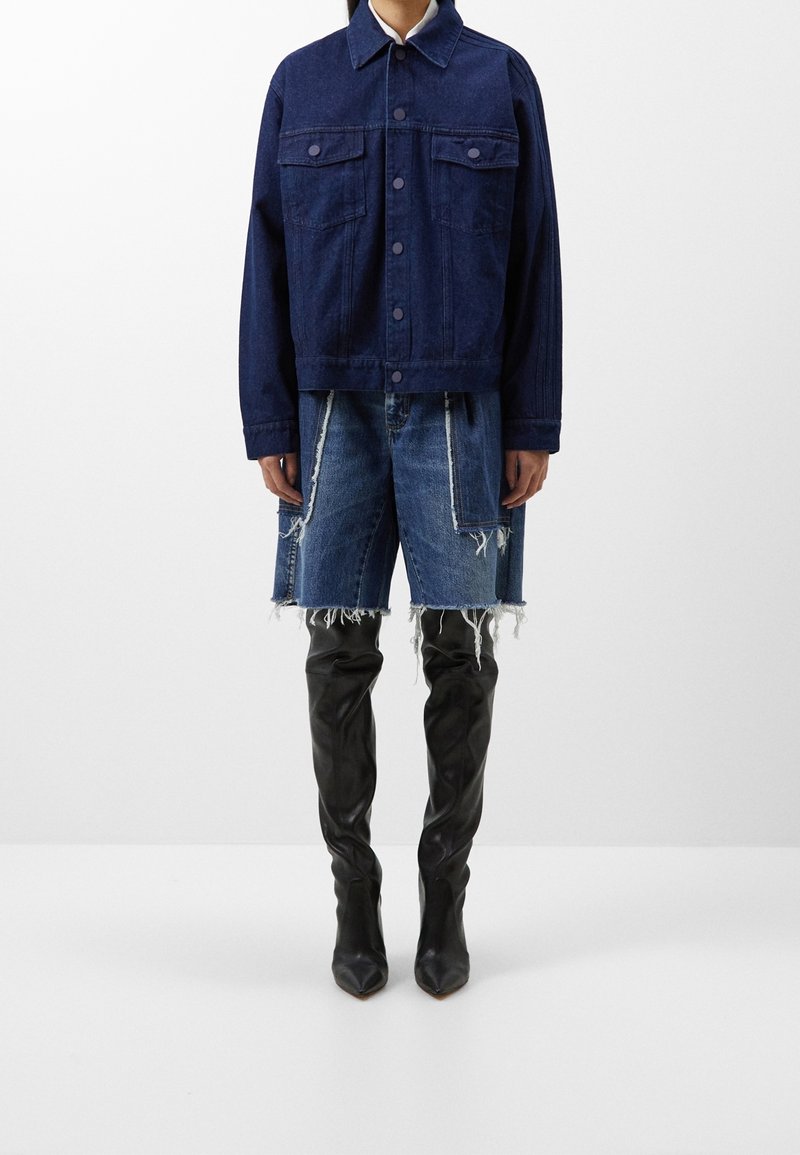 Laivastonsininen farkkutakki napillaisella suljennalla, kahdella rintataskulla, yhdistettynä kulutettuihin sinisiin farkkushortseihin ja korkeisiin mustiin piikkikärkisiin saappaisiin.