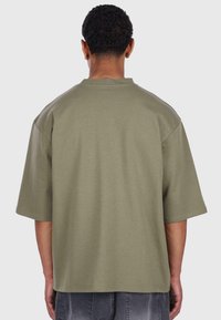 T-shirt oversize verde oliva, maniche corte, scollo rotondo, texture morbida e orlo dritto. Dettaglio della cucitura posteriore visibile.