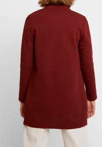 Veste tunique en tissu texturé bordeaux, avec manches longues, col rond et coupe droite s'étendant sous les hanches.