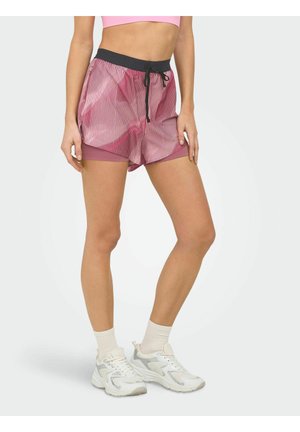 Vrouw in roze gestreepte sportshorts met interne compressielaag, witte sneakers en witte sokken, staand tegen een effen achtergrond.