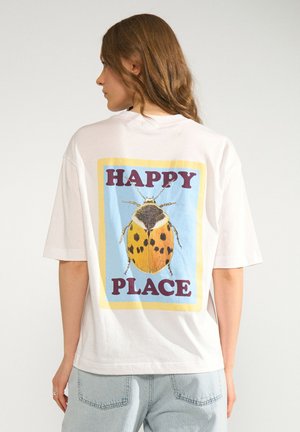Femme aux longs cheveux bruns portant un t-shirt blanc avec une grande coccinelle jaune et noire et les mots "HAPPY PLACE" dans le dos.