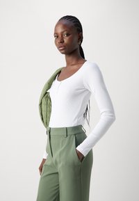 White ribbed long-sleeve top with a scoop neckline, paired with high-waisted, light green tailored pants, and a green quilted jacket draped over the shoulder.
