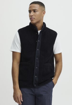 Zwarte, pluche fleece vest met een hoge kraag, knoopsluiting en twee voorzakken. Gemaakt om te dragen over een wit shirt, met een relaxed fit.