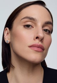 Okrągłe, metalowe kolczyki w kolorze szarym i czarnym, połączone z gładką skórą i naturalnym makijażem modelki na jasnym tle.