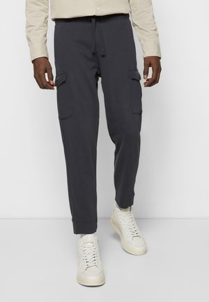Mann trägt dunkelgraue Cargo-Hose mit Klappentaschen und weißen High-Top-Sneakers, steht vor weißem Hintergrund.