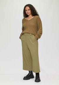 Bruine gebreide trui met V-hals, gecombineerd met olijfgroene broek met wijde pijpen met knoopaccenten en voorzakken. Zwarte veterschoenen.