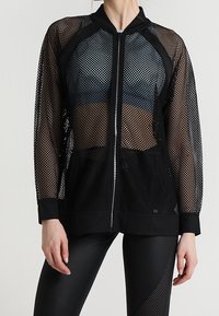 Woman wearing black mesh zip-up jacket over sports bra and black leggings with mesh detail, standing against plain light background.