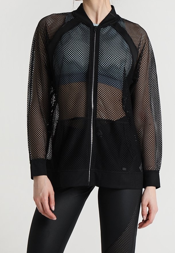 Woman wearing black mesh zip-up jacket over sports bra and black leggings with mesh detail, standing against plain light background.