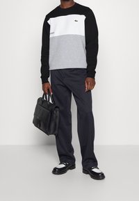 Uomo che indossa un sweatshirt Lacoste a blocchi di colore, pantaloni neri, scarpe in bianco e nero, mentre tiene una valigetta in pelle nera su uno sfondo bianco.