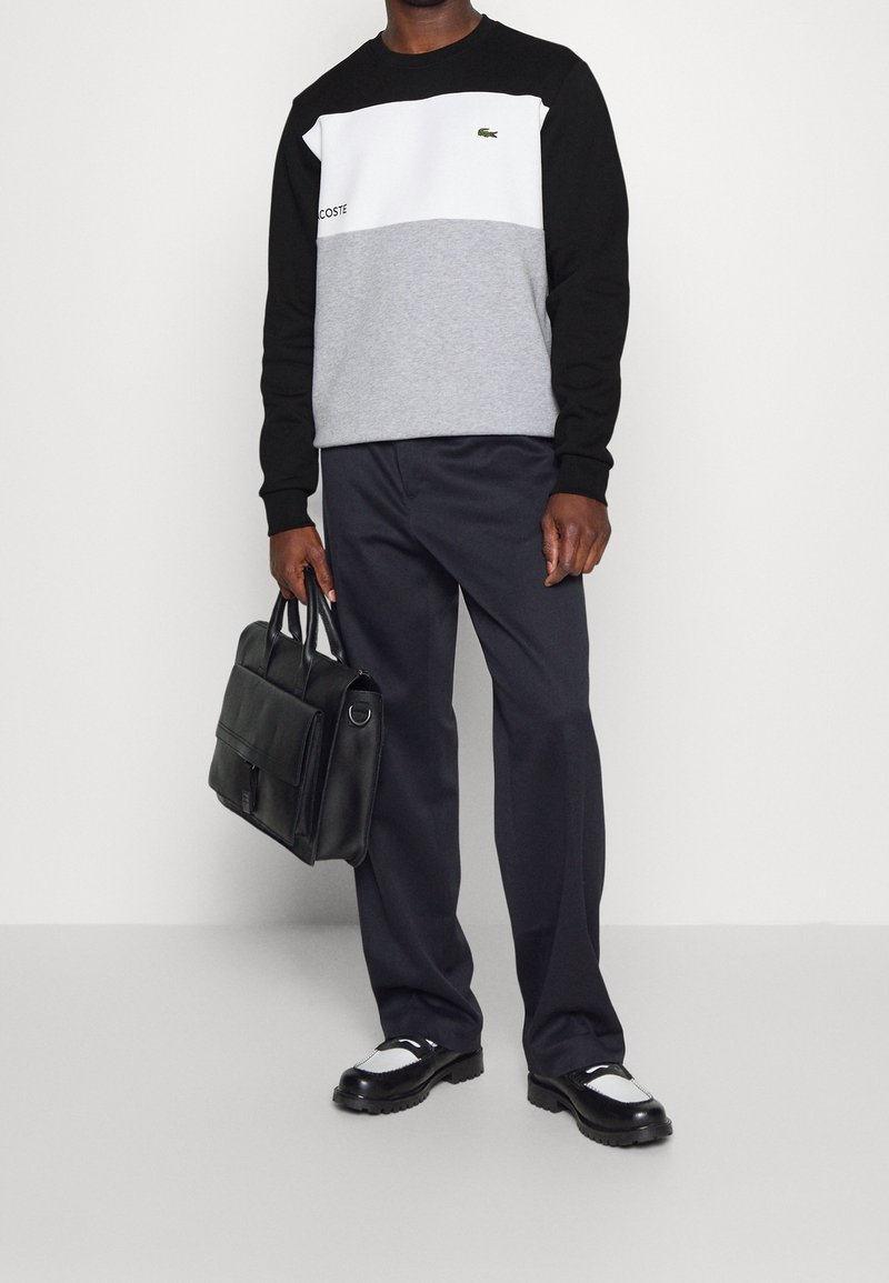 Uomo che indossa un sweatshirt Lacoste a blocchi di colore, pantaloni neri, scarpe in bianco e nero, mentre tiene una valigetta in pelle nera su uno sfondo bianco.