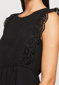 Vestido negro con detalles de encaje ornamentado en los hombros, textura suave de la tela y detalle fruncido en la cintura. Escote clásico redondo.