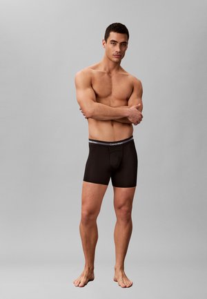 Shirtless man with muscular build wearing black Calvin Klein boxer briefs, standing barefoot against plain gray background with arms crossed.