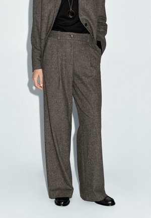 Frau trägt hoch taillierte, weit geschnittene braune Tweed-Hose mit schwarzen Schuhen und einem schwarzen Oberteil, eine Hand in der Tasche und eine sichtbare Anhängerkette.