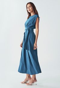 Zila, bezroku maksi kleita ar teksturētu audumu, jostu viduklī un vaļēju piegriezumu, kopā ar baltām stiletto sandālēm.