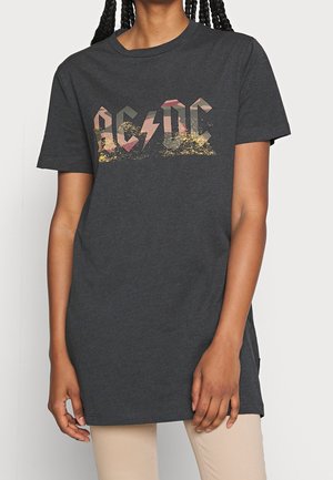 Osoba nosząca ciemnoszarą koszulkę z wyblakłym wielokolorowym logo AC/DC, w połączeniu z jasnobeżowymi spodniami, stojąca na jednolitym tle.