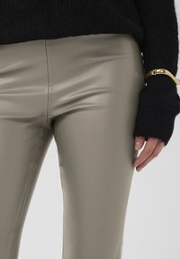 Pantalon beige en similicuir avec une finition élégante, associé à un pull en maille noir et un bracelet manchette doré porté sur un poignet partiellement visible.