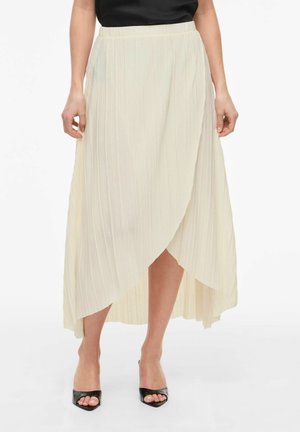 Sieviete valkā krēmkrāsas krokas apkārt apģērbu ar asimetrisku apakšmalu un melnas, atvērtu purngalu papēžu sandales.