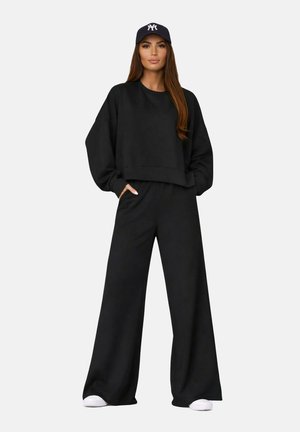 Femme portant un sweat-shirt noir ample et un pantalon à jambes larges, des baskets blanches et une casquette de baseball bleu marine avec un logo blanc, debout les mains dans les poches.