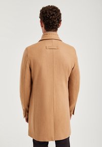 Kappa i tan, sydd av en ullblandning med en ensam bakslits, snedkrage och en rektangulär knappdetalj vid halsen; figursydd passform och långa ärmar.