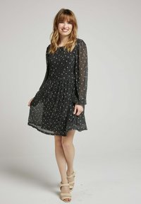 Robe noire, longueur genou, avec de longues manches transparentes et un tissu texturé. Présente une jupe plissée et des accents dorés dispersés. Portée avec des talons beiges.