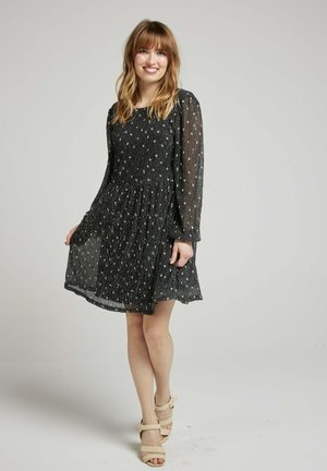 Robe noire, longueur genou, avec de longues manches transparentes et un tissu texturé. Présente une jupe plissée et des accents dorés dispersés. Portée avec des talons beiges.