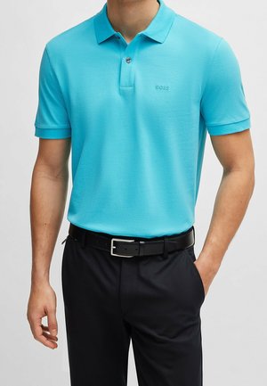 Mann trägt ein türkisfarbenes Kurzarm-Polo mit „BOSS“-Logo, in schwarze Hose mit schwarzem Gürtel gesteckt, Hand in der Tasche.