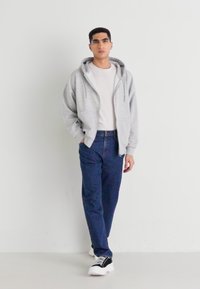 Joven caminando, vestido con una sudadera gris claro, camiseta blanca, jeans azul oscuro y zapatillas negras y blancas, contra un fondo blanco liso.