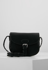 Černá kožená crossbody taška s předním klopou zabezpečenou stříbrnou sponou, nastavitelné řemínky stočené vedle ní na bílém povrchu.