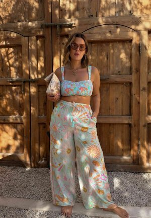 Frau trägt ein pastellfarbenes, sternmusterndes Crop-Top und weite Hosen, hält eine Blumen-Clutch und steht barfuß vor Holztüren.