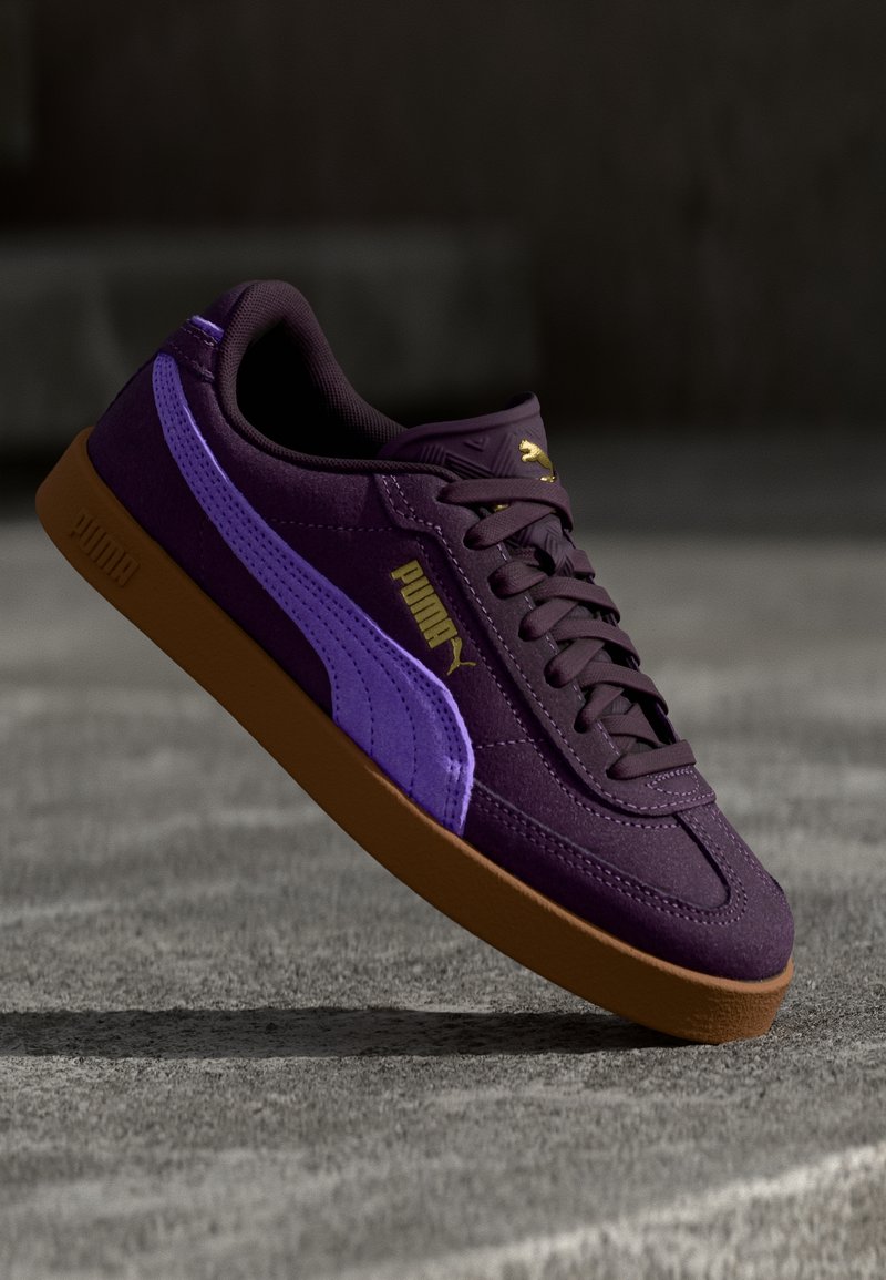 Fioletowe zamszowe sneakersy z grubą gumową podeszwą, z jasnofioletowym paskiem, złotym logo i zaokrąglonym noskiem.