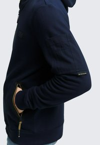 Person in einem navyblauen Kapuzenpullover mit einem Reißverschluss-Seitenfach und einer Stofftasche am Oberarm, Markenetikett auf dem Ärmel sichtbar.
