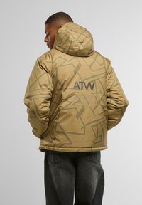 Giacca puffer beige con un motivo geometrico, dotata di cappuccio e polsini elasticizzati. Il retro mostra il logo "ATW" in grigio. Indossata sopra pantaloni grigio scuro.
