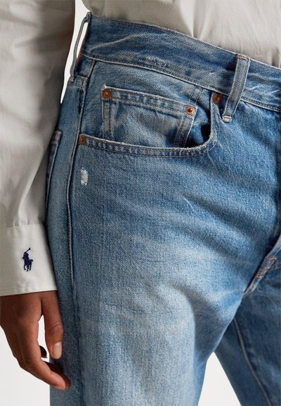 Inimese lähivõte, kes kannab helesiniseid teksapükse ja valget pika varrukaga särki, mille mansetil on tumesinise tikandiga logo.