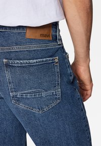 Jeans de mezclilla azul oscuro con una etiqueta de cuero marrón que dice "mavi" en la cintura trasera, y un bolsillo trasero con costuras detalladas.