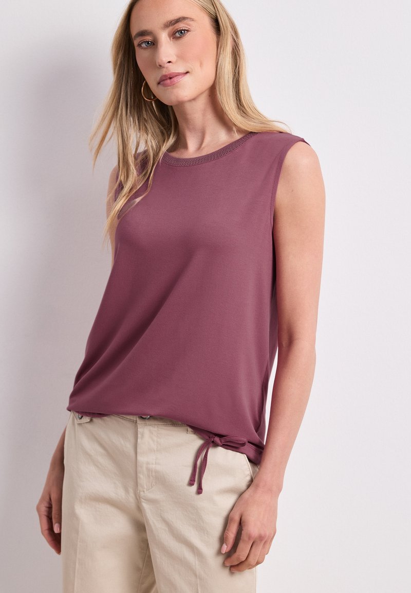 Femme aux longs cheveux blonds portant des boucles d’oreilles créoles dorées, un top mauve sans manches avec un lien sur le côté, et un pantalon beige sur un fond clair uni.