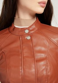 Veste en cuir marron avec un col haut en tricot beige côtelé, accentuée par deux boutons-pression argentés et de subtils détails de couture.