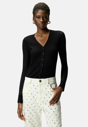 Donna che indossa un cardigan nero a coste con bottoni e pantaloni bianchi decorati con una griglia di piccole borchie d'argento, posando con una mano in tasca.