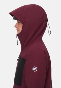 Bordeauxfärgad huva jacka med en svart bröstficka, justerbara dragsko och en vit logotyp på ärmen. Slät, väderbeständig tyg.