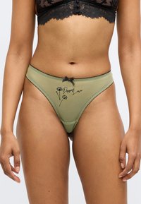 String vert en mesh avec un imprimé floral noir et le texte "Poppy Love". Présente un petit nœud noir et une bordure décorative autour de la taille.