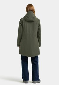 Olivgrön vattentät jacka med huva, långa ärmar och en något utfläkt fåll, i kombination med mörkblå jeans och grå skor.