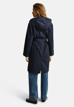 Femme portant un manteau ceinturé à capuche bleu marine, manches jaune clair visibles, jean large bleu et chaussures foncées, debout de dos.