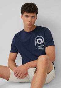 Navyblaue Baumwoll-T-Shirt mit kreisförmigem weißen Grafik, das eine Blume und Text zeigt. Kurze Ärmel und runder Halsausschnitt, kombiniert mit hellbeigen Shorts.