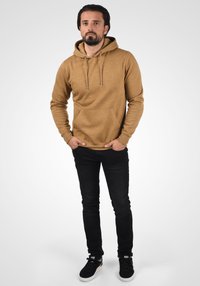 Brun hættetrøje med en frontlomme og snore, parret med sorte jeans og sorte sneakers, stående mod en lys gradient baggrund.