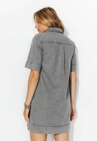 Femme aux cheveux blonds portant une robe chemise en denim gris à manches courtes, tournée pour montrer le dos, devant un fond blanc uni.