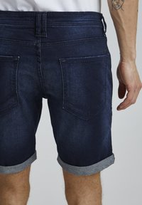 Pantalones cortos de mezclilla azul oscuro con dobladillos enrollados, que cuentan con dos bolsillos traseros y detalles cosidos. El material es suave con un ligero brillo.