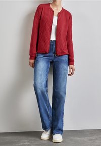 Chaqueta bomber acolchada roja con cremallera frontal, cuello redondo y dos bolsillos laterales. Combinada con jeans azul claros y zapatillas blancas.