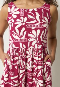 Vestido sin mangas con grandes patrones florales blancos y abstractos sobre un fondo rosa intenso, llevado con las manos en los bolsillos.