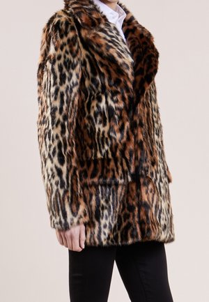 Personne portant un manteau en fausse fourrure à imprimé léopard sur une chemise blanche et un pantalon noir, debout devant un fond clair uni.