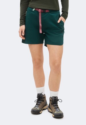 Άτομο που φοράει σκούρα πράσινα σορτς με ροζ ζώνη, λευκές κάλτσες και καφέ ορειβατικά μποτάκια, στέκεται με το ένα χέρι στην τσέπη.