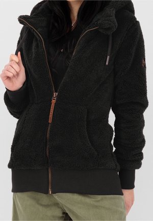 Musta vetoketjullinen fleece-takki hupulla ja etutaskuilla, pidetty mustan topin päällä ja yhdistetty oliivinvihreisiin housuihin.